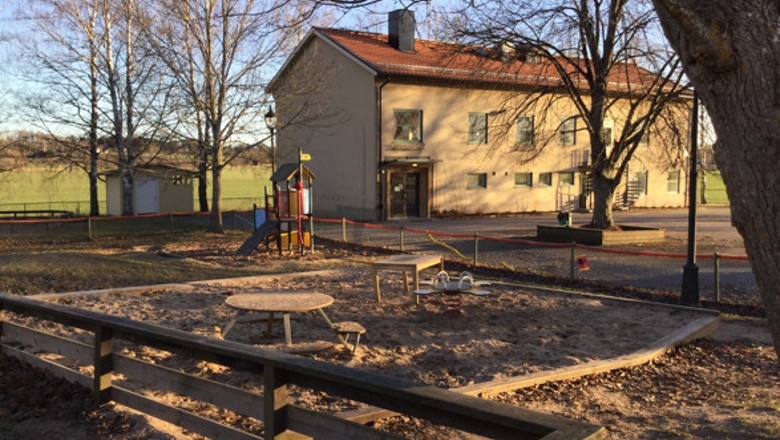 I förgrunden ett staket, sandlåda med olika leksaker i. I bakgrunden ett äldre tvåvåningshus med tegeltak.i