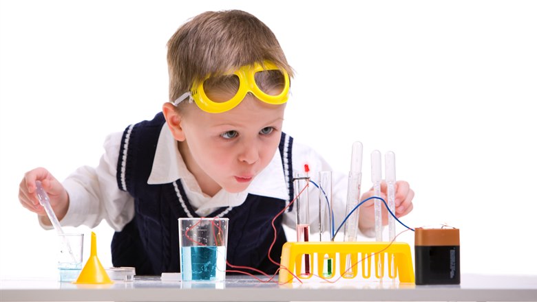 En liten pojke med gula glasögon i pannan håller på med ett experiment med batteri, sladdar och vätska