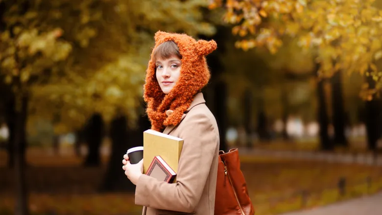 En kvinna som håller i en bok och en kaffemugg. Hon har en stickad mössa i form av en räv på sig.
