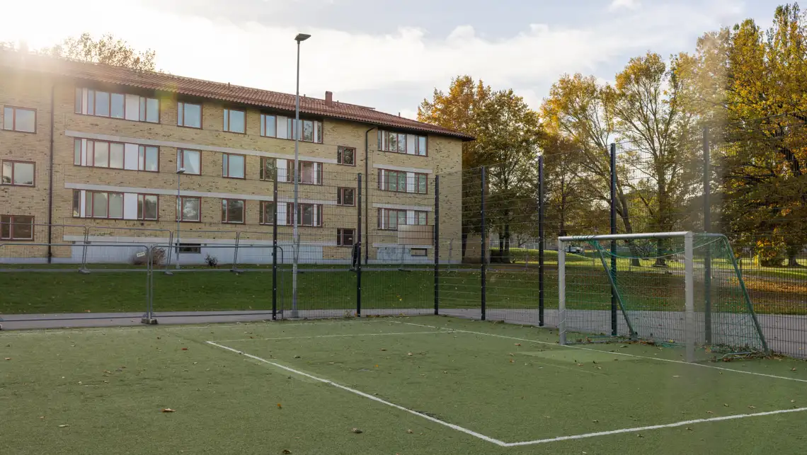I förgrunden syns en gammal fotbollsplan. I bakgrunden syns ett hyreshus i Berga