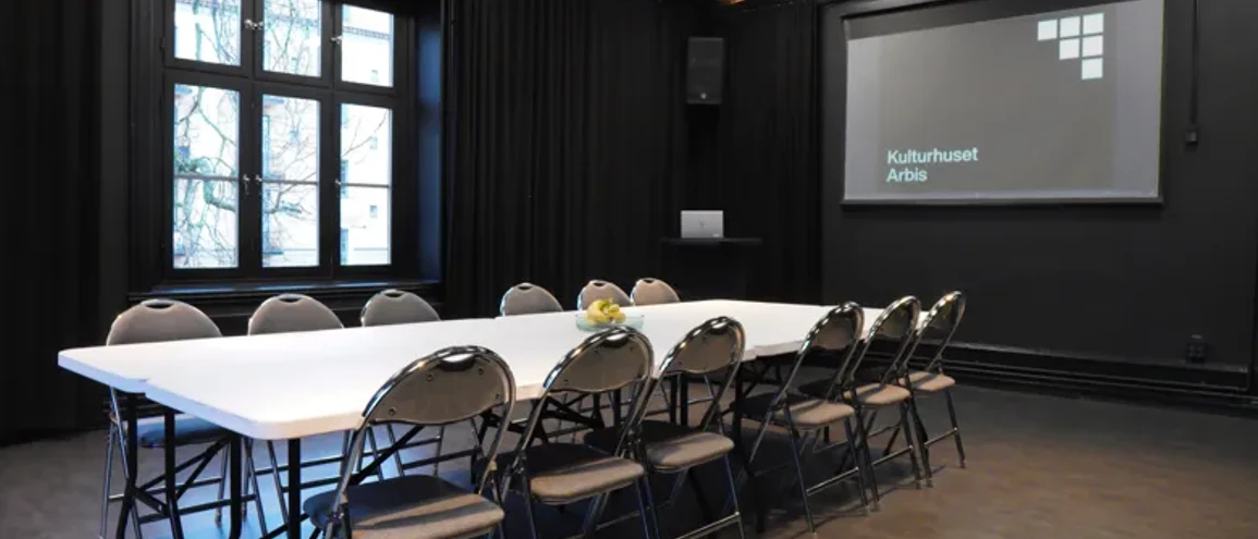 Konferenslokal med vitt bord omgivet at tio klappstolar och en storbildsskärm