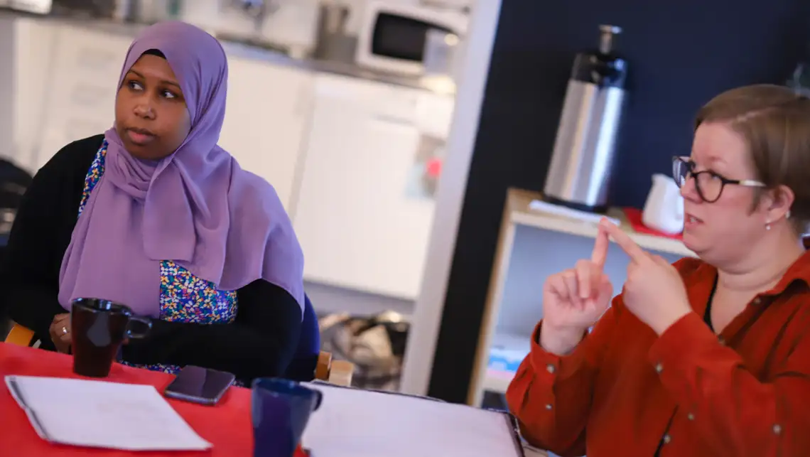 Frida Edler håller upp sina händer medan hon pratar. Hon bär en röd skjorta. Bredvid henne sitter en kvinna.