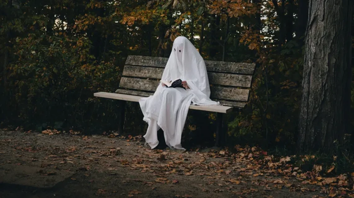 En person utklädd till spöke med ett vitt lakan sitter på en parkbänk och läser en bok