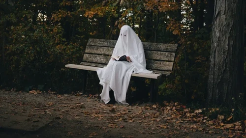En person utklädd till spöke med ett vitt lakan sitter på en parkbänk och läser en bok