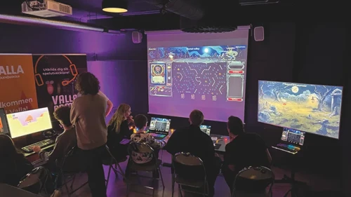 Ett antal personer sitter och tittar på storbildsskärmar  i ett nedsläckt rum
