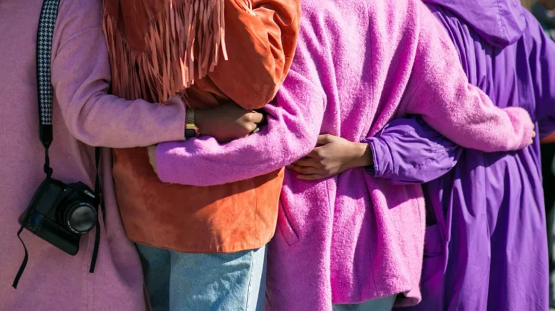 Ryggarna av fyra personer i lila, rosa och orange kläder, som håller varandra om ryggen