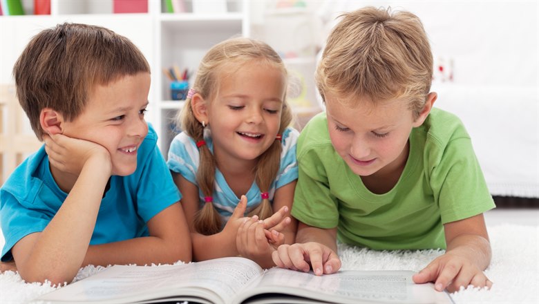 tre leende barn, två killar och en tjejm,  ligger på mage på en matta och läser i en bok tillsammans