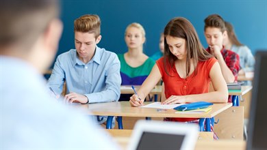 Elever sitter vid bänkar i ett klassrum och skriver. I förgrunden en lärare med en iPad.