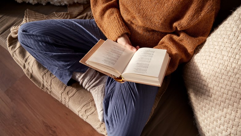 En kvinna i en varm tröja sitter i en soffa och läser en bok.