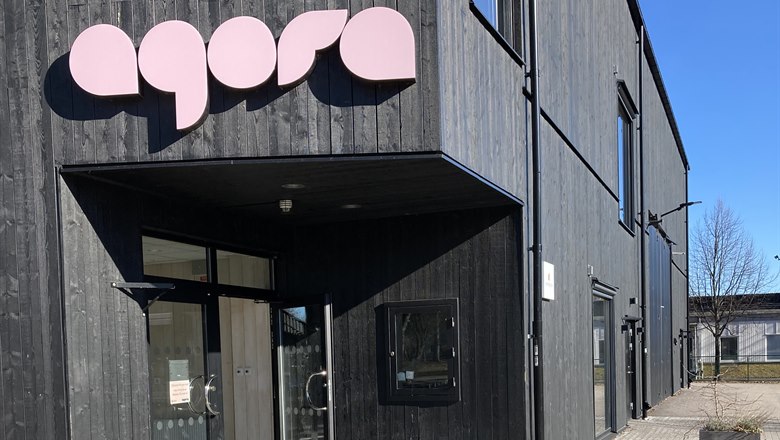 Agorahuset. En byggnad i svart trä med texten Agora i rosa över ingången. En dörr står öppen och framför ingången står en trottoarpratare.