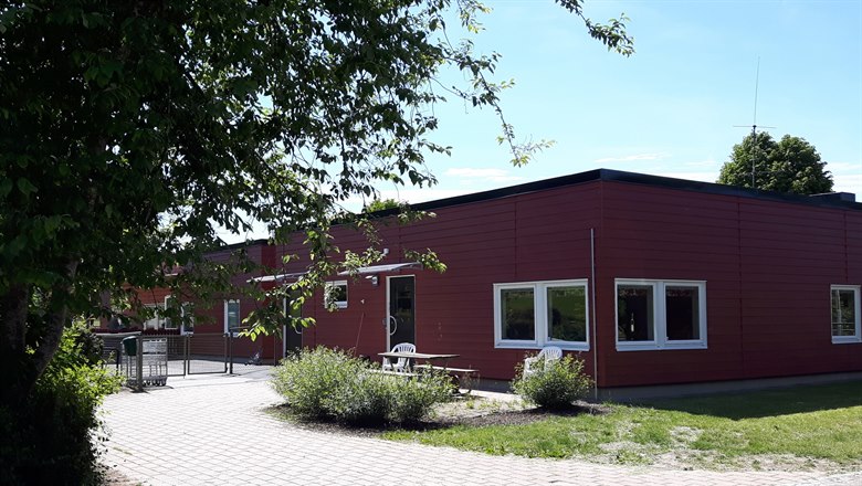 En röd förskolebyggnad inbäddad i gröna buskar och gräsytor, en stengång fram till huset.