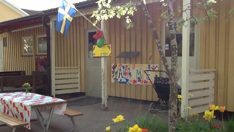 Entré tillförkloa med blommor ien rabatt, med ett bord och svenska flaggan , det finns en färgglad skylt som det står välkommen på
