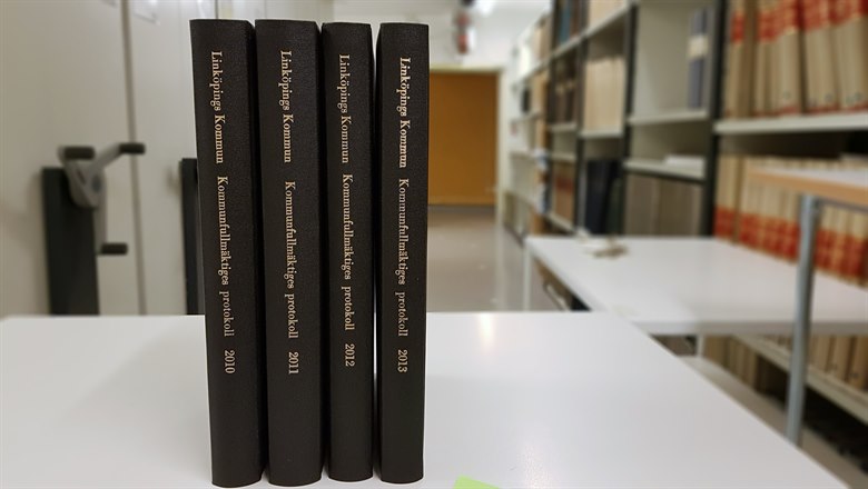 Fyra svarta böcker med kommunfullmäktigeprotokoll, i bakgrunden arkivhyllor