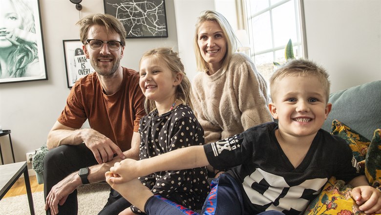 En familj med två föräldrar och två barn sitter i en soffa i sitt vardagsrummet. Sonen i familjen är närmast kameran och ler in i kameran. 