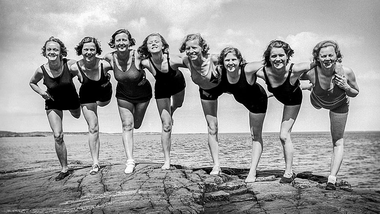 Svartvitt. Åtta yngre kvinnor står i baddräkt på ett ben på klippsten vid en sjö.