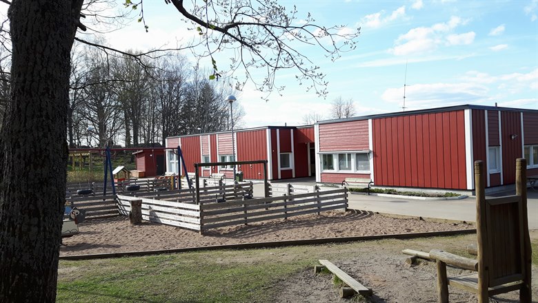 Vy över delar av lekgården med en trätron i förgrunden och förskolans röda hus i bakgrunden