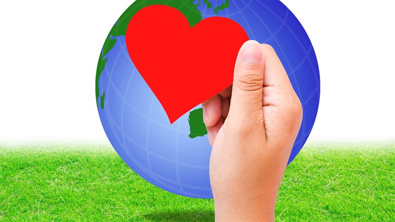 En hand håller upp ett rött pappershjärta framför en jordglob med grönt gräs under