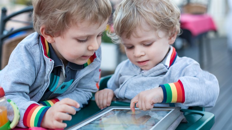 Två små pojkar sitter vid ett bord och använder en iPad. De har likadana jackor och lockigt hår.