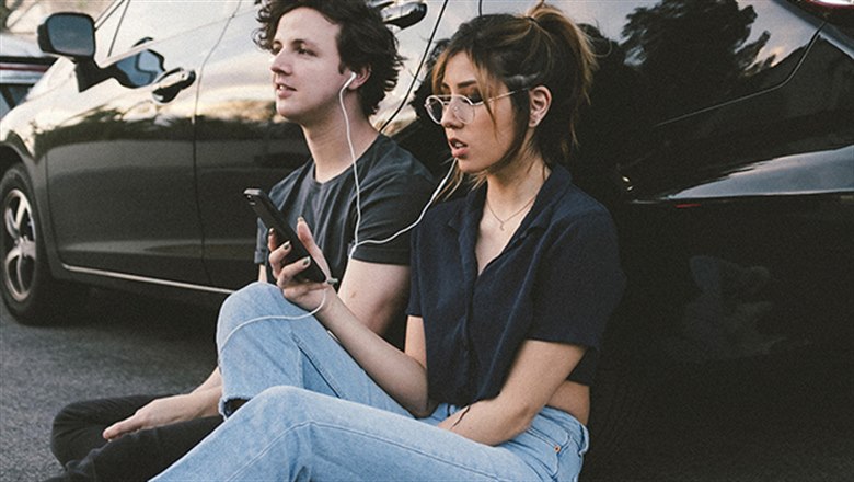 Två ungdomar sitter lutandes mot en bil och lyssnar på musik genom varsin hörlur.