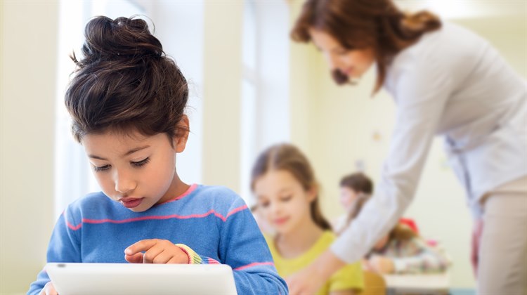 En flicka sitter i ett klassrum och använder en iPad. Bakomh henne sitter fler barn och en lärare står lutat över flickan bakom