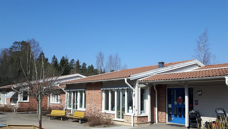 Framsidan på förskolan Bronsåldersgatan 50. Långt tegelhus med blå dörr och gula bänkar framför.