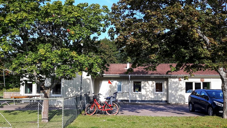 Förskolans ljusgula hus sett från Älgvägen. En röd cykel och en svart bil står parkerad framför, några lummiga träd i förgrunden.