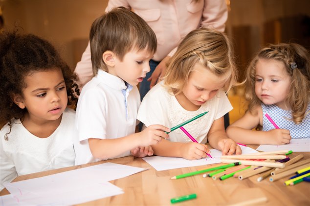 Fyra förskolebarn sitter med tuschpennor och papper tillsammans. I bakgrunden en pedagog.